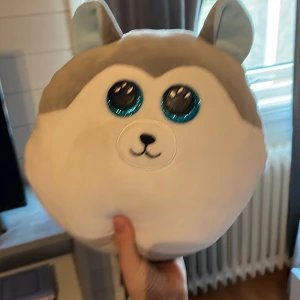 Mjukisdjur Squishmallows Husky - Supermjuk och rund Squishmallows husky från Ty med stora glittriga blå ögon, grå och vit päls samt små öron. Perfekt som kramkompis eller dekoration. Märkt med 'Slush' på etiketten.