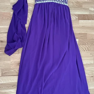 Lila långklänning med pärldetaljer - Elegant lila långklänning med glittriga pärldetaljer upptill och avtagbar sjal. Klänningen har en luftig och svepande passform, perfekt för speciella tillfällen. Resår i ryggen för extra komfort.