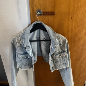 Croppad ljusblå jeansjacka med slitningar - Säljer en croppad jeansjacka i ljusblå denim med slitna detaljer och råa kanter. Jackan har två bröstfickor med knappar och klassisk krage. Perfekt för en trendig och avslappnad stil.