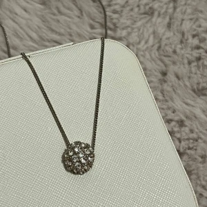 Silverfärgat halsband med glittrig kula - Säljer ett elegant silverfärgat halsband med en rund, glittrig hängande kula täckt av små stenar. Perfekt för att ge lite extra bling till din outfit. Kedjan är tunn och stilren.