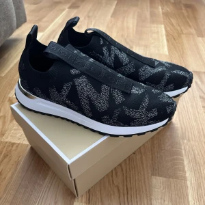 Svarta slip-on sneakers från Michael Kors - svarta slip-on sneakers från Michael Kors med silvriga detaljer på hälen och mönstrad ovandel. Skorna har en vit och svart sula med gummidobbar undertill och elastiskt band över foten med logga. Perfekta för en sportig och trendig look.