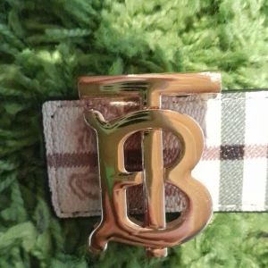 Burberry-inspirerat bälte med guldspänne - Säljer ett snyggt bälte med klassiskt rutigt mönster i beige, svart och vitt. Bältet har ett stort guldigt spänne med initialerna 'TB'. Perfekt accessoar för att lyfta din outfit. Den är inte använt helt perfekt double sided