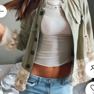 Grön jacka med virkade detaljer - Snygg ljusgrön jacka med stora svarta knappar och virkade, krämvita detaljer med tofsar vid ärmslut och nederkant. Jackan har två bröstfickor och en rak passform. Perfekt för dig som vill sticka ut med en unik stil.