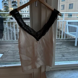 Vit linnetopp med svart spets - Säljer en vit linnetopp med djup v-ringning och breda svarta spetsdetaljer längs ringningen. Toppen är ärmlös och har en mjuk, glansig yta som ger en lyxig känsla. Ny pris 500. Köpt på MQ. Måste strykas lite. Skriv om ni är intresserade!