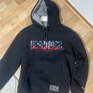 Svart hoodie från Hoodrich - Svart hoodie från Hoodrich med stor logga i svart, vitt och rött på både fram- och baksidan. Tröjan har huva med dragsko, magficka och ribbade muddar. Detaljerad patch nertill och broderad text på ärmen.