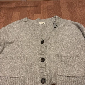 Ljusgrå stickad kofta med stora knappar - Mysig ljusgrå stickad kofta med stora svarta knappar och två framfickor. Koftan har långa ärmar och bred ribbstickad mudd vid ärmslut och nederkant. Perfekt för lager-på-lager-stil.