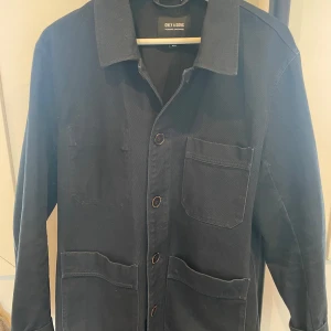 Blå overshirt från ONLY & SONS - Säljer en blå overshirt från ONLY & SONS i storlek M. Jackan har klassisk krage, knappar framtill och tre stora fickor på framsidan. Perfekt att slänga över en t-shirt för en clean look.