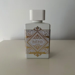 Lattafa Badee Al Oud Honor & Glory 80-90ml - Parfym i matt vit färg med geometriska guldmönster och texten 'Badee Al Oud Honor & Glory' på framsidan. Flaskan har en fyrkantig form och ett vitt lock med gulddetalj på toppen. Märket Lattafa är ingraverat på baksidan. Cirka 80-90ml kvar! 