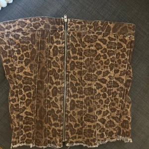 Leopardmönstrad tubtopp med dragkedja - Säljer en snygg tubtopp med leopardmönster och dragkedja framtill. Toppen har råa kanter nertill och smock baktill för en bekväm passform. Perfekt för dig som vill sticka ut med ett djurmönster!