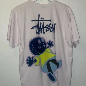 Stussy doll tee - Säljer en vit t-shirt från Stüssy med stort grafiskt tryck på ryggen och mindre tryck på bröstet. T-shirten har normal passform och är tillverkad i mjuk bomull. Perfekt för dig som gillar streetwear och unika prints. Kvitto finns om du behöver🙏🏻