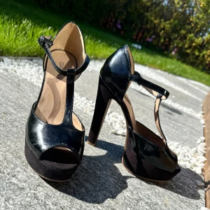 Svarta högklackade pumps med T-rem  - Säljer ett par svarta pumps från Catwalk med hög klack och platåsula. Skorna har T-rem med spänne runt vristen och öppen tå. Glansigt material upptill och mockaliknande platå och klack. Perfekta för dig som vill sticka ut.