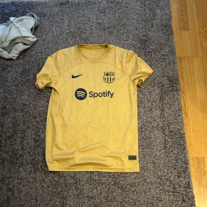FC Barcelona bortatröja Lewandowski 9 - Säljer en gul FC Barcelona bortatröja från Nike med Lewandowski 9 på ryggen. Tröjan har korta ärmar med regnbågsfärgade detaljer och sponsorloggor från Spotify och UNHCR. Perfekt för dig som älskar Barca och vill sticka ut på läktaren.
