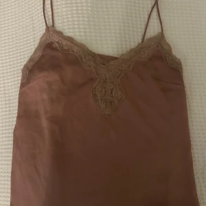 Rosa linnetopp med spets från Bik Bok - Säljer en ljusrosa linnetopp från Bik Bok med tunna axelband och vacker spetsdetalj längs urringningen. Toppen har en mjuk, glansig yta och är perfekt för lager-på-lager eller sommarens varma dagar.