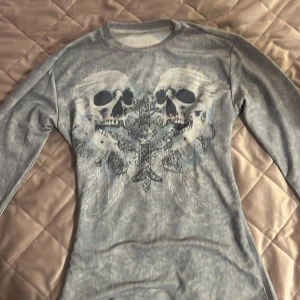 Grå långärmad tröja med dödskalletryck - Säljer en grå långärmad tröja med ribbad struktur och stort tryck av två dödskallar och ett detaljerat kors framtill. Tröjan har rund halsringning och är figurnära i modellen. Perfekt för dig som gillar edgy stil.