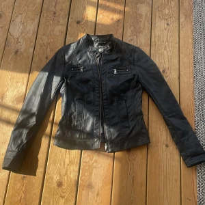 Svart skinnjacka med dragkedja - Snygg svart skinnjacka med ståkrage och dragkedja framtill. Jackan har två bröstfickor med dragkedja och markerade sömmar för en stilren look. 