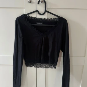 Svart croppad topp med spets från Even&Odd - Säljer en svart långärmad croppad topp från Even&Odd med spetsdetaljer vid halsringning och nederkant. Toppen har v-ringning och är perfekt för att skapa en snygg och trendig look.