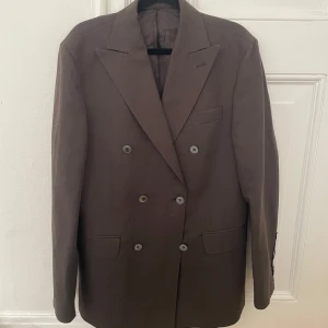 Brun dubbelknäppt kavaj från Oscar Jacobson - Säljer en brun dubbelknäppt kavaj från Oscar Jacobson. Klassisk modell med breda slag, fyra knappar framtill och tre knappar vid ärmslutet. Fodrad insida med märkets logga. Perfekt för dig som vill ha en stilren och tidlös look. Har använt endast 1 gång