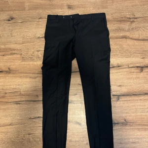 Svarta kostymbyxor från Riley Shelby - Snygga svarta kostymbyxor från Riley Shelby. Klassisk rak passform med hällor för bälte och diskreta bakfickor med knappar. Perfekta för en stilren look.