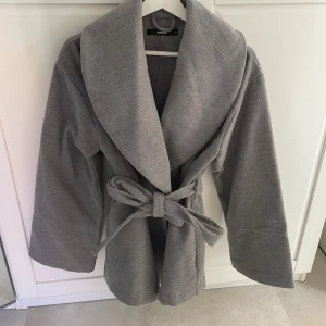 Grå kappa med bälte från Bik Bok - Säljer en grå kappa från Bik Bok med bred krage och knytskärp i midjan. Jackan har lång ärm och en avslappnad passform, perfekt för lager-på-lager. Enkel och stilren design som passar till många outfits.