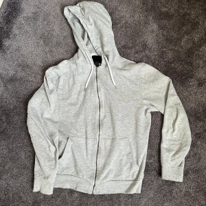 Ljusgrå hoodie med dragkedja - Säljer en ljusgrå hoodie från 157 med dragkedja framtill och huva med snören. Tröjan har långa ärmar och två fickor framtill. Perfekt för en avslappnad stil.