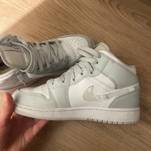 Nike Air Jordans Mid i vitt och ljusgrått - Säljer ett par Nike Air Jordans Mid i vitt och ljusgrått skinn med klassisk swoosh-logga på sidan. Skorna har snörning, perforerad tå och platt sula. Perfekta för dig som gillar stilrena sneakers med ikonisk design.