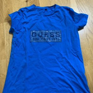 Blå t-shirt från Guess med strassdetaljer - Säljer en blå t-shirt från Guess med glittrande strassdetaljer på bröstet där det står 'GUESS SINCE EST 1981'. T-shirten har korta ärmar och en klassisk rund halsringning. Perfekt för dig som gillar lite extra bling i vardagen.