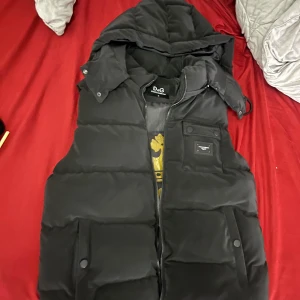 Svart dunväst från D&G med huva - Säljer en svart dunväst från D&G med huva och dragkedja. Västen har två stora fickor framtill med knappar och en innerficka. Insidan har ett stort D&G-tryck i guld med krona. Perfekt för lager-på-lager-stil.