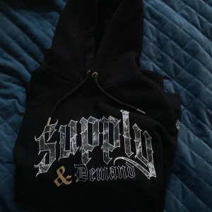 Svart hoodie från Supply & Demand - Svart hoodie från Supply & Demand med stor vit och guldig text på bröstet. Klassisk huva med dragsko och mjukt material. Perfekt för en streetwear-look och riktigt skön att bära. Köpt i England! Tröjan är i storlek XS men passar även S.