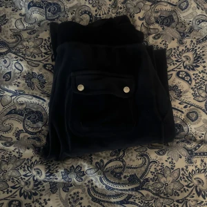 Svarta velourbyxor från Juicy Couture - Mysiga svarta velourbyxor från Juicy Couture med fickor framtill som har silverfärgade knappar. Byxorna har en klassisk passform och är perfekta för en avslappnad stil.