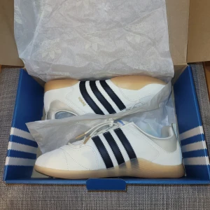 Adidas Samba x BadBunny - Säljer ett par Adidas Samba OG sneakers i vitt skinn med klassiska svarta ränder och gummisula. Skorna har spänningssnörning och låg profil, perfekt för en sportig och stilren look. Ikonisk modell med svart detalj på plösen. Vill tillägga att storleken är egentligen 39 ¹/3 men passar säkert någon med storlek 39.
