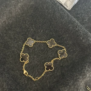 Guldfärgat armband med svarta fyrklöver - Säljer ett elegant armband med fem svarta fyrklöver i guldfärgad kedja. Armbandet har en klassisk och stilren design som passar perfekt till att lyfta din outfit. Lås med karbinhake. Tål vatten 
