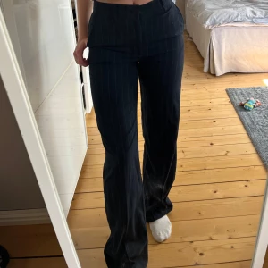 Svarta/ marinblåa kostymbyxor med vita ränder från Pull&Bear - Snygga mörkblåa/svarta kostymbyxor från Pull&Bear med tunna vita ränder. Byxorna har normal passform, raka ben och klassisk midja. Perfekta för en stilren look med fickor i sidorna och knappstängning framtill.