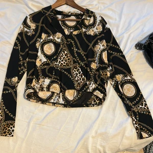 Svart leopardmönstrad blus Gina Tricot - Snygg svart blus från Gina Tricot med leopardmönster och kedjedetaljer i guld och beige. Blusen har lång ärm, klassisk krage och omlott-design framtill. Materialet är mjukt och följsamt, perfekt för dig som gillar att sticka ut med coola prints.