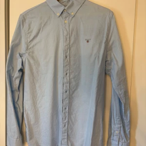 Ljusblå skjorta från GANT - Snygg ljusblå skjorta från GANT med klassisk krage och knappar hela vägen. Diskret logga broderad på bröstet. Skjortan har lång ärm och är gjord i mjuk bomull som känns skön mot huden. Perfekt för dig som gillar stilrena och tidlösa plagg. Str 16 year old 176cm passar som S. Aldrig använd bara tvättad