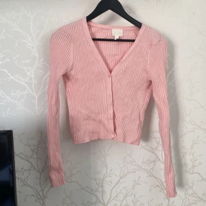 Rosa ribbad kofta från H&M - Supersöt ljusrosa kofta från H&M i ribbad bomull. Modellen är croppad med v-ringning och knappar framtill. Långärmad och perfekt för dig som gillar en chill och feminin stil. Passar till jeans eller kjol för en snygg look.
