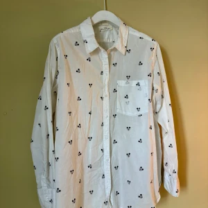 Vit skjorta med Mickey Mouse-mönster - Säljer en vit skjorta från H&M med svarta Mickey Mouse-huvuden som mönster över hela plagget. Skjortan har klassisk krage, bröstficka och långa ärmar med knappar vid manschetterna. 