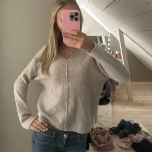 Beige stickad v-ringad tröja - Mysig beige stickad tröja med v-ringning och synlig söm framtill. Modellen är loose fit med långa ärmar och mjukt material som känns skönt mot huden. Perfekt för chill dagar och enkel att matcha med jeans eller kjol.