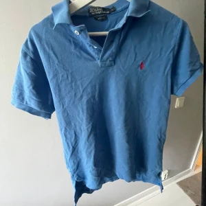 Blå Polo Ralph Lauren piké S - Blå t-shirt från Polo Ralph Lauren i storlek S, klassisk pikémodell med krage och två knappar. Röd broderad logga på bröstet. Tillverkad i mjuk bomull och har korta ärmar. Custom fit för en snygg passform.