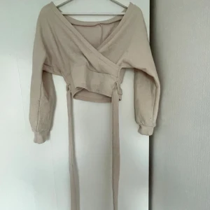 Beige omlottopp med långa band - Säljer en beige croppad omlottopp med långa knytband i midjan och v-ringad hals. Toppen har långa ärmar med muddar och är gjord i ett mjukt bomullsmaterial.  