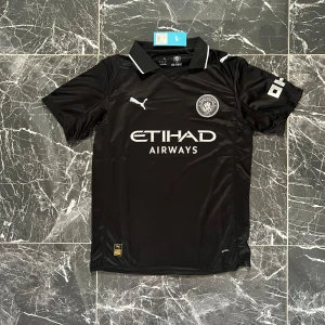 Manchester City svart fotbollströja Puma - Snygg svart Manchester City fotbollströja från Puma med vit logga och detaljer. Tröjan har klassisk krage med vit kant, korta ärmar och tryckta sponsorloggor på bröstet och ärmen. Tillverkad i lätt, ventilerande polyester som passar perfekt på planen.