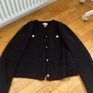 Svart kofta med guldknappar - Snygg svart kofta med guldknappar framtill och på fickorna. Koftan har rund hals, ribbade muddar och är stickad i ett mjukt material. Perfekt för dig som gillar klassisk stil med en twist. Fyra fickor framtill ger extra detaljer.