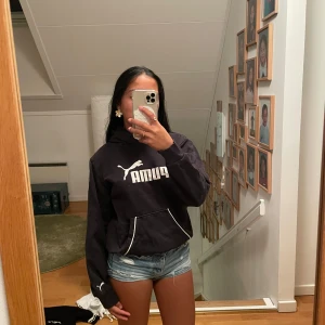 Svart hoodie från Puma med logga - Svart hoodie från Puma med stor vit logga på bröstet och huva. Tröjan har en klassisk känguruficka med vita detaljer och långa ärmar. Mjuk och skön bomullsmix, perfekt för chill eller sport. Snygg streetstil som funkar året runt.