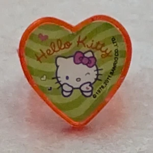 Hello Kitty hjärtring i plast - Supergullig ring i orange plast med ett hjärtformat motiv av Hello Kitty. Ringen har gröna och ljusgröna ränder, lila och rosa detaljer samt texten 'Hello Kitty' i orange. Perfekt för dig som älskar Hello Kitty och vill ha något unikt på fingret! Ringen har en mindre defekt på plast hjärtat visas på bild.