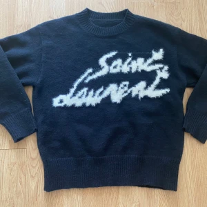 Svart Saint Laurent stickad tröja - Svart stickad tröja från Saint Laurent med vit, fluffig logga framtill. Tröjan har rund halsringning, ribbade muddar och är långärmad. Perfekt för dig som vill ha en statement piece med lyxig känsla och cool design.