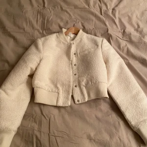 Vit teddyjacka med knappar - Säljer en vit teddyjacka med croppad passform och ribbade muddar. Jackan har rund hals och stängs med snygga knappar framtill. Perfekt för dig som gillar mjuka och fluffiga material med en modern twist.