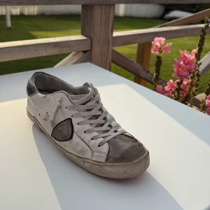 Philippe Model vita sneakers med grå detaljer - Snygga sneakers från Philippe Model i vitt skinn med grå mockadetaljer på tå och sidor. Mörkblå häl med orange kant, och coolt märke på sidan. Yttersulan är orange och har ett mönstrat grepp. 