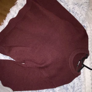 Vinröd stickad tröja från Ginatricot - Mysig vinröd stickad tröja från Ginatricot med rund hals och långa ärmar. Tröjan har en enkel och clean design med ribbade muddar vid hals, ärmslut och nederkant. Perfekt för att hålla sig varm under kyliga dagar.