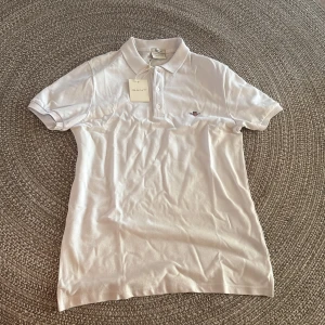 Vit pikétröja från GANT - Fräsch vit pikétröja från GANT med klassisk krage och korta ärmar. Tröjan har broderad GANT-logga på bröstet och knäppning med tre vita knappar. Materialet är mjuk bomull som andas och känns skönt mot huden. Perfekt för en clean och stilren look.