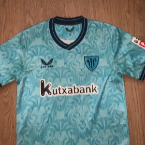 Blå Athletic Club Bilbao fotbollströja - Säljer en snygg blå Athletic Club Bilbao fotbollströja från Castore med coolt abstrakt mönster. Kortärmad med mörkblå detaljer vid krage och ärmslut. Materialet är lätt och andas, perfekt för match eller träning. Stor logga på bröstet och sponsortryck.