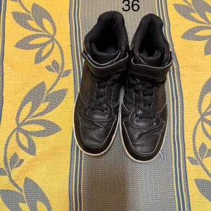 Svarta höga sneakers med snörning - Vi säljer en svart höst- och vinterkänga i storlek 36. Den har snörning framtill och kardborreband upptill för extra stöd. Yttermaterialet är tillverkat av konstläder, innermaterialet är fodrat för värme och sulan är vit med ett grovt mönster undertill.